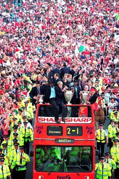 a large group of people standing on top of a double decker bus in front of a crowd