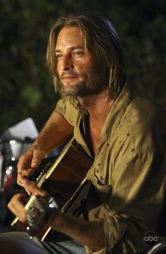 a man with long hair sitting on a motorcycle holding an acoustic guitar and looking at the camera