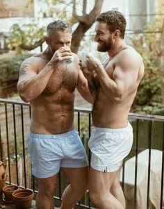 two men standing next to each other on a balcony with no shirts and one holding a glass in his hand