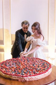 a man and woman cutting into a large cake on top of a wooden table next to each other