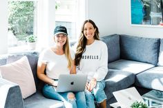 two women sitting on a couch with a laptop computer in front of them, both smiling at the camera