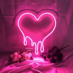 a pink neon heart shaped sign sitting on top of a bed next to dried flowers