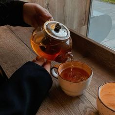 a person pouring tea into two mugs on a wooden table next to a window