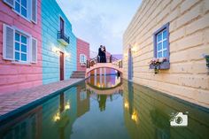 a woman standing on a bridge over a small pond in front of colorful buildings with windows