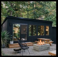 a small black cabin with a fire pit and picnic table in the back yard, surrounded by trees