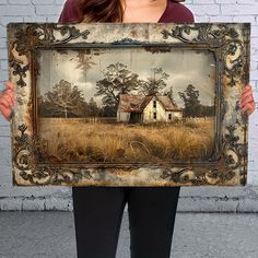 a woman holding up a painting in front of a brick wall with an old house on it