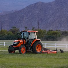 an orange tractor is plowing the grass in front of a white fence and mountains