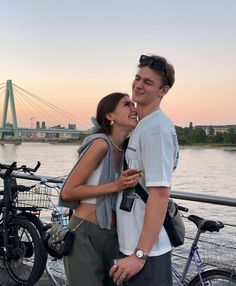 a young man and woman standing next to each other near water with bicycles in the background