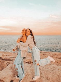 two women hugging each other on top of a rock next to the ocean at sunset