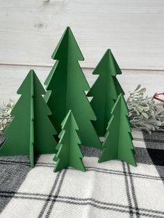 three green paper christmas trees sitting on top of a plaid tablecloth next to an evergreen branch