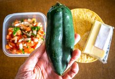 a hand holding a green pickle next to a container of food