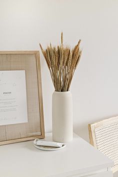 a white vase with dry grass in it next to a framed photograph and pen on a table