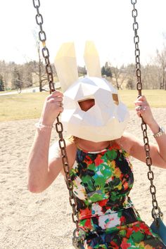 a woman sitting on a swing wearing a paper bunny mask over her face and head