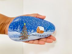 a hand holding a painted rock with a christmas tree on it and a house in the background