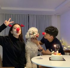 two people with clown noses and nose paint holding a dog in front of a birthday cake