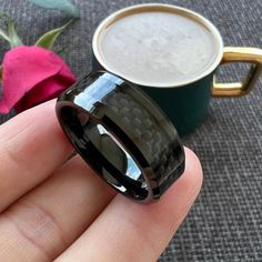 a hand holding a black ring next to a cup of coffee and pink rose on the table