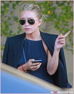 a woman in sunglasses is holding her cell phone and pointing to the side with one hand