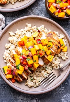 two plates with chicken, rice and mango salsa on them next to a bowl of grilled vegetables