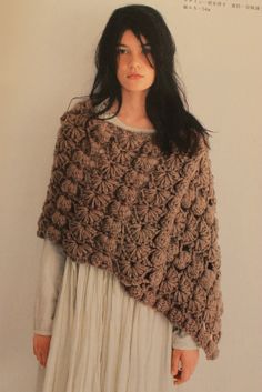 a woman wearing a brown crochet shawl standing in front of a white wall