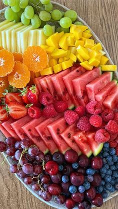 a platter filled with different types of fruit on top of a wooden table next to oranges, raspberries, grapes and watermelon