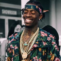 a man with tattoos wearing a flowery shirt and gold chain around his neck smiles at the camera