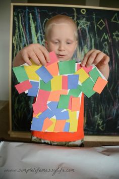 a young boy holding up a piece of paper with colored squares on it, in front of a blackboard