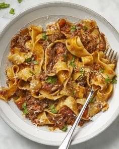 a white bowl filled with pasta and meat on top of a marble table next to a fork