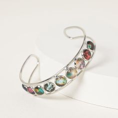 a silver bracelet with multicolored stones on the outside and inside, sitting on a white surface