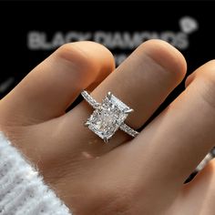 a woman's hand with a diamond ring on top of her finger, in front of some bushes