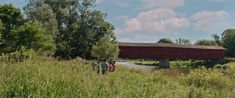 two people and their dogs are walking in the grass near a covered bridge with a red roof