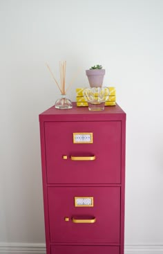 a pink filing cabinet with gold handles and drawers on top, next to a potted plant