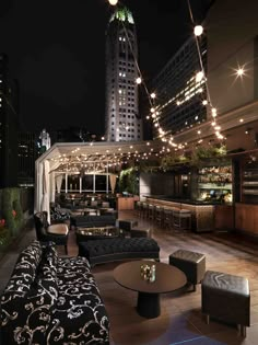 an outdoor seating area at night with lights strung from the building's roof and high rise buildings in the background