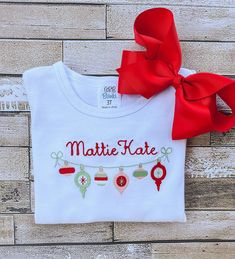 This adorable girl's shirt is perfect for Christmas and would be a super cute outfit for a Christmas card! The detailed design of the Christmas Ornaments, as well as your chosen name, is created by machine embroidery and uses high quality thread.  The white shirt is true to size and is made out of 100% combined cotton interlock fabric to keep your child completely comfortable. The shirt has naturally tapered shoulders and includes a ruffled hemline. The short sleeve shirts have puff sleeves.  Pl Holiday Embroidered Tops As Gift, Embroidered Christmas Holiday Tops, Christmas White Embroidered Tops, White Tops With Custom Embroidery For Christmas, Cute White Top For Festive Occasions, White Christmas Tops With Embroidered Graphics, Gift Red Embroidered Top, Red Embroidered Holiday Tops, Red Embroidered Tops Gift