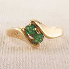 This vintage two stone ring captures two emeralds in a 14K Yellow Gold waved band. Each Emerald measures roughly 3.3mm with one emerald having a slight chip as seen in pictures. The classic and simple styling gives this ring a timeless elegance. This ring is currently a size 6 1/4 US. Total Weight: 4.4g This ring can be sized between US sizes 4-9. For custom sizing of this ring please contact us for pricing. Resizing will add additional time to delivery. A custom listing will be created to inclu Timeless Emerald Ring For Anniversary, Vintage Emerald Ring For Promise, Classic Tsavorite Emerald Ring With Prong Setting, Fine Jewelry Emerald Ring With Tension Setting, Classic 14k Gold Emerald Ring With Tension Setting, Timeless Green Emerald Ring Stamped 14k, Formal Emerald Ring With Tension Setting For May Birthstone, Green Emerald Three Stone Ring In 14k Gold, Three Stone Emerald Ring In 14k Gold