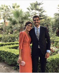 a man and woman standing next to each other in front of some bushes with trees