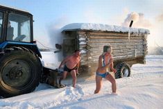 two men in swimsuits are standing next to a tractor on the snow covered ground