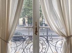 an open window with white curtains and a wrought iron fence in front of it, looking out onto the street