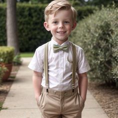 a young boy wearing a bow tie and suspenders standing on a sidewalk in front of bushes