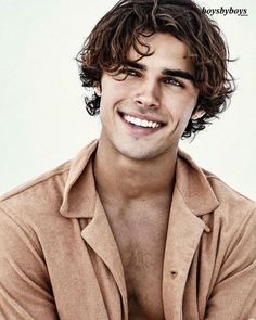 a young man with curly hair smiling at the camera while wearing a tan shirt and brown pants