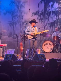 two men on stage playing guitars and singing into microphones with other musicians in the background