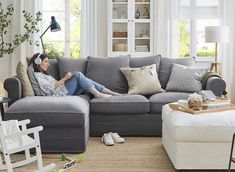 a woman sitting on top of a gray couch in a living room next to a window