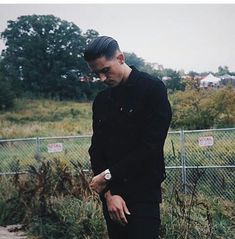 a man standing in front of a fence looking at his cell phone while wearing all black