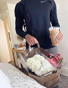 a man standing next to a bed holding a cup of coffee and flowers in his hand