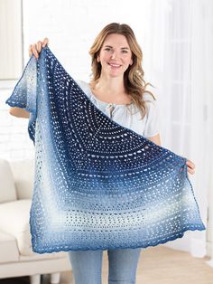 a woman holding up a blue crocheted shawl in front of a white couch