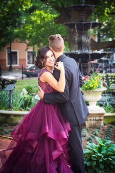 a man in a tuxedo hugging a woman in a purple dress and standing next to a fountain