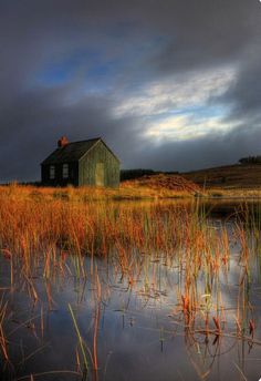 an old house sitting on top of a field next to a body of water with tall grass in front of it