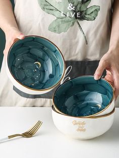 two bowls with gold rims are being held by a woman's hands while another bowl holds a fork and knife