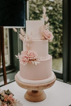 a three tiered wedding cake with pink flowers and feathers on the top is sitting on a table