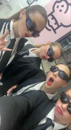 three women in suits and sunglasses posing for the camera with one pointing at her phone