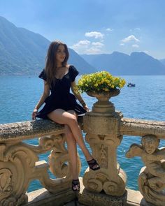 a woman sitting on top of a stone fence next to the ocean with flowers in her lap